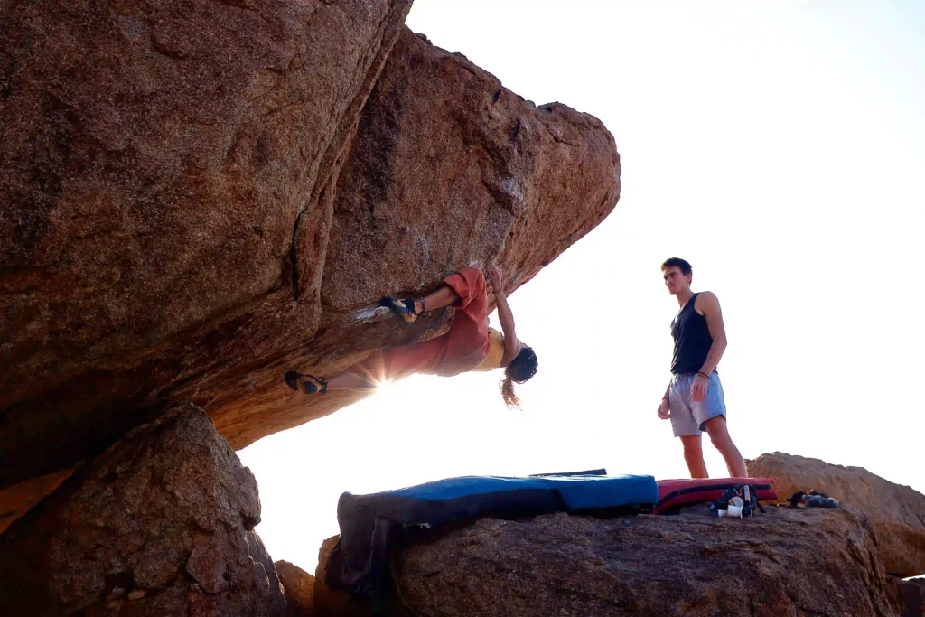 A woman bouldering upside down on overhang outdoors boulder in Perth WA, with man supporting and looking on.