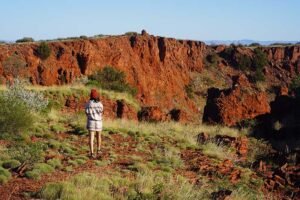 Female hiker with red beanie at peak of Mount Bruce in Karijini National Park with blue skies and red ochre rock face in background.