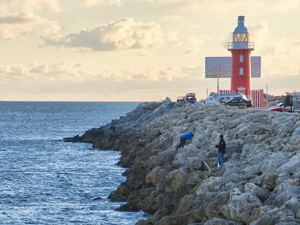 North Mole, WA fishing spot in Fremantle with several fishers on rocks next to water and red lighthouse in background.