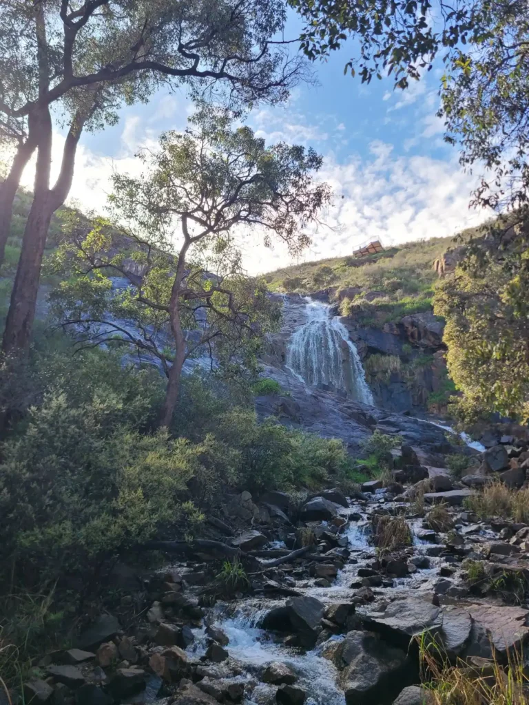View from foot of Lesmurdie Falls in Perth Hills, with hiking trail on right and stream collecting at photographer's foot on a blue sky day.