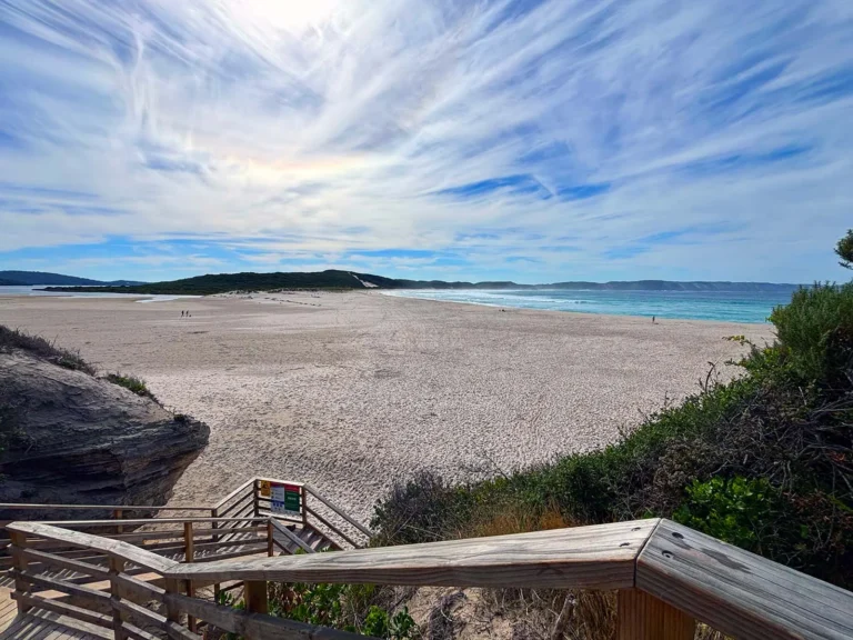 Landscape photo of ocean beach in denmark wa from stairs toward beach, blue sky and clouds above with footsteps on sand
