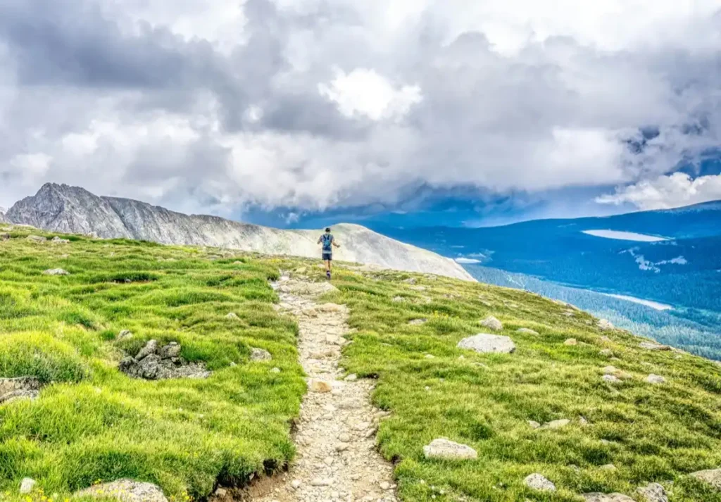Trail runner in middle of path leading over a grassy knoll with mountains and grey clouds in background