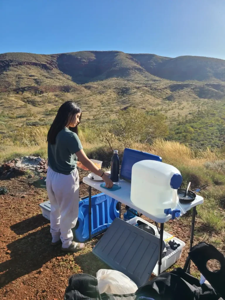 Female camper preparing breakfast on fold out table overlooking the Hamersely Range at Albert Tognolini Rest Area in Karijini National Park, WA.
