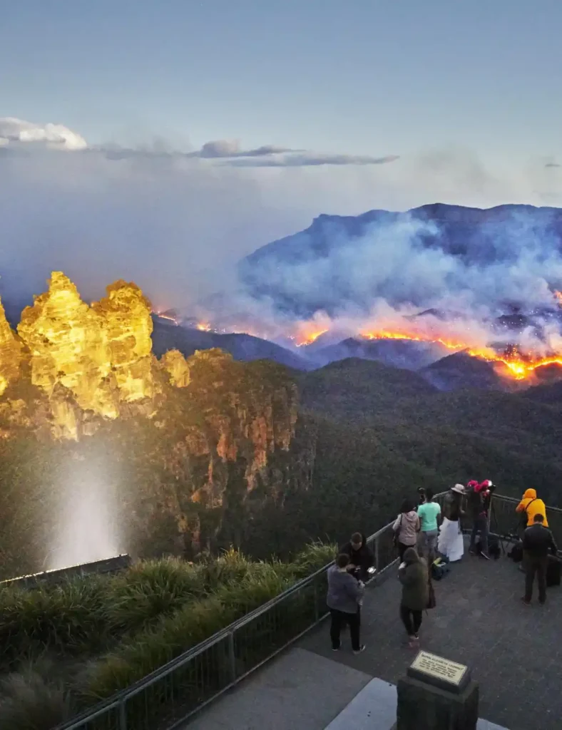 The three sisters in the Blue Mountains lit up in yellow lights at dusk while a fire roars across a valley of forest. View is from the lookout with multiple tourists overlooking the flames.