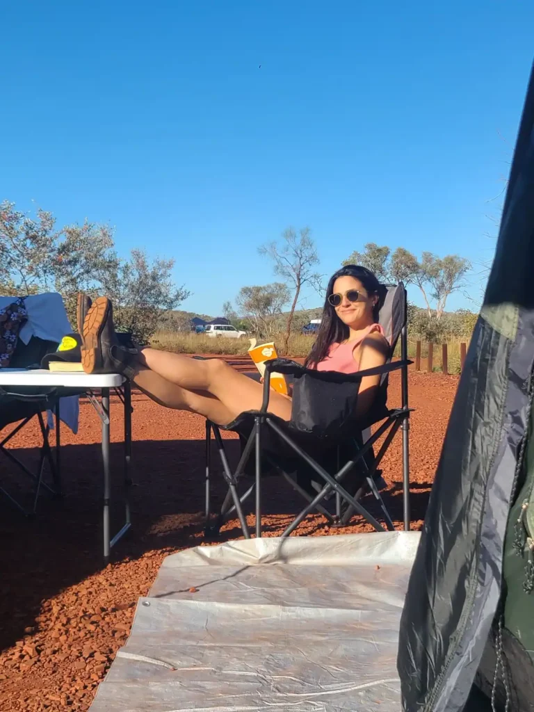 Female hiker with feet up on fold out table at dales campground near Karijini National Park reading a book with blue sky above.