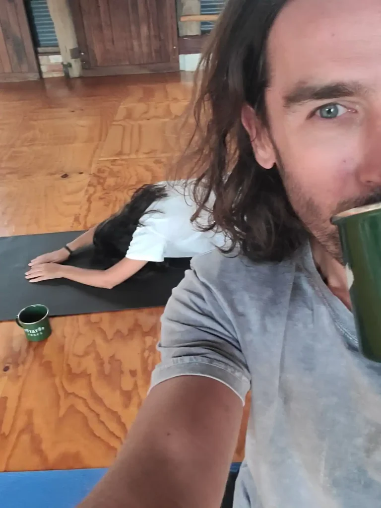Woman doing yoga at Fair Harvest campground with male camper in foreground sipping tin cup of coffee.