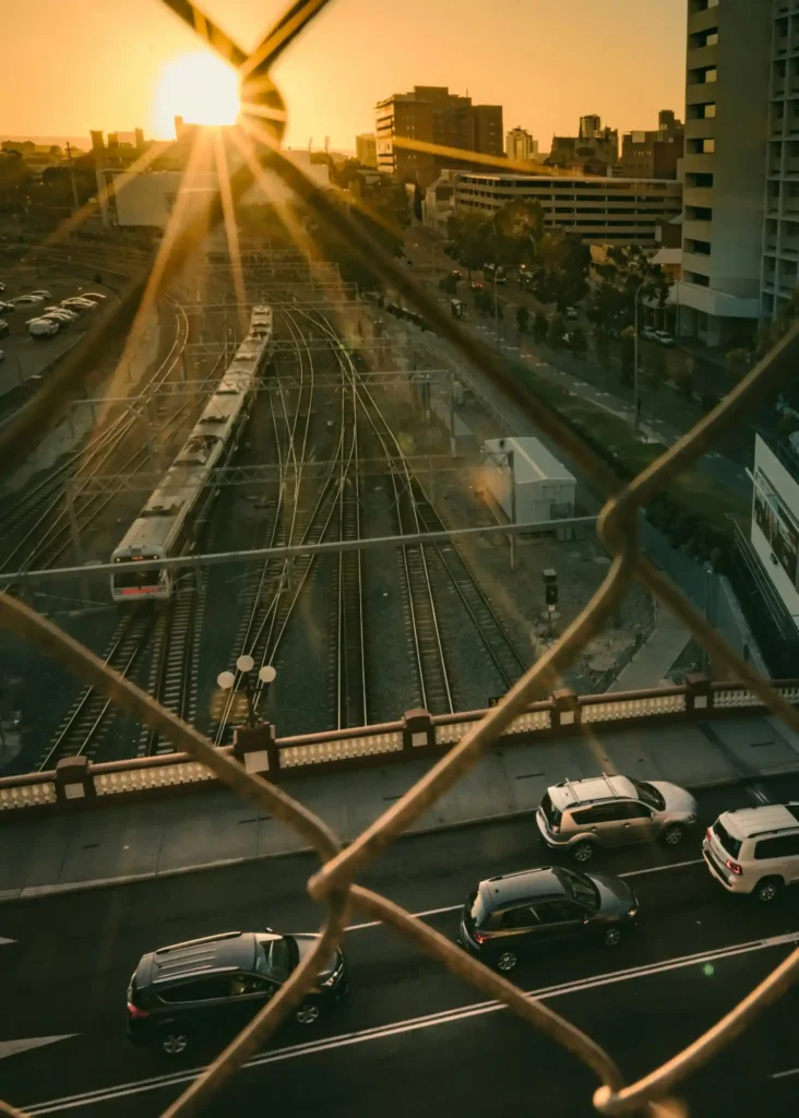 Image of sun rising above train in Perth train station, with congested freeway traffic in foreground. Photo taken behind steel gate, with orange sky illuminating the early city morning.
